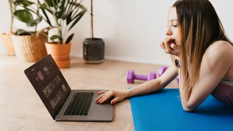 A woman on an exercise mat choosing a workout on her laptop