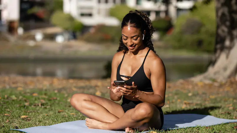 Woman sitting on a yoga mat outdoors checking her phone