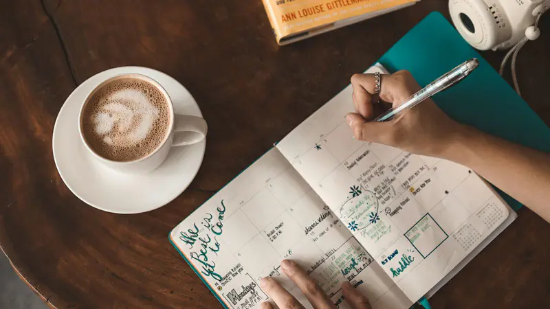 A woman writing in a planner beside a cup of coffee
