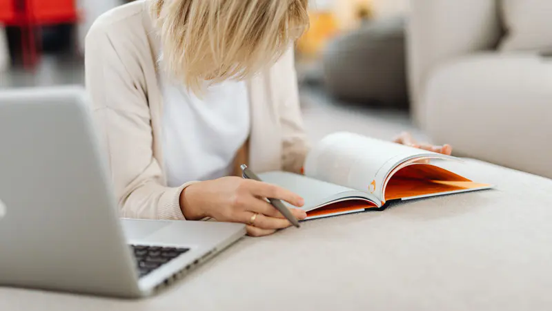A woman reading a book with her laptop open beside her