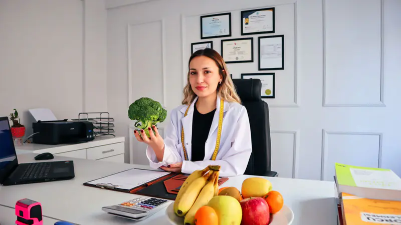 A nutritionist at her desk holding broccoli, with fruit in front of her