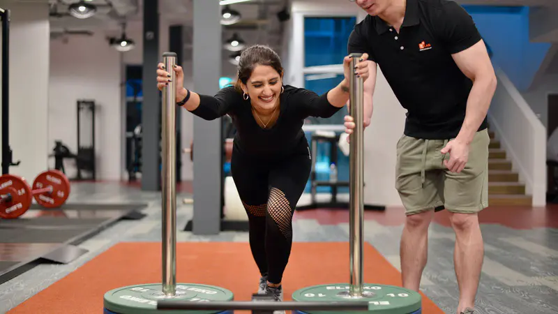Woman pushing a weighted sled in a gym with a coach beside her