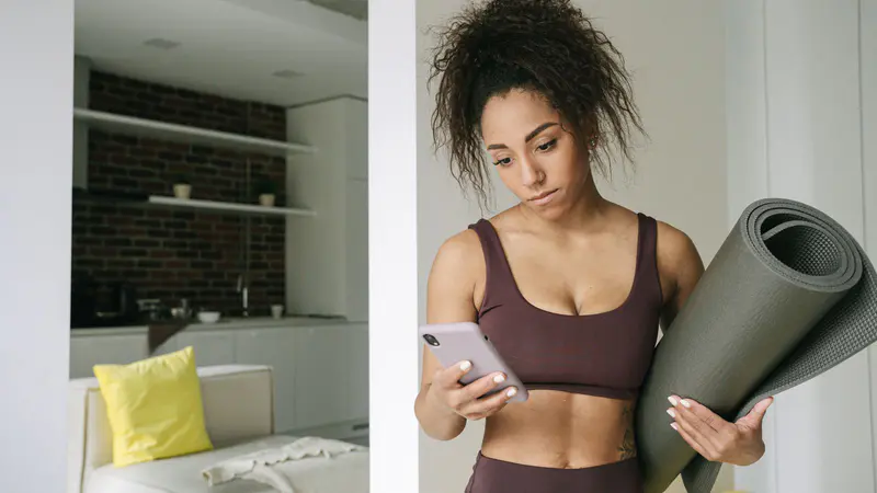 A woman in activewear checking her phone, with a yoga mat under her arm